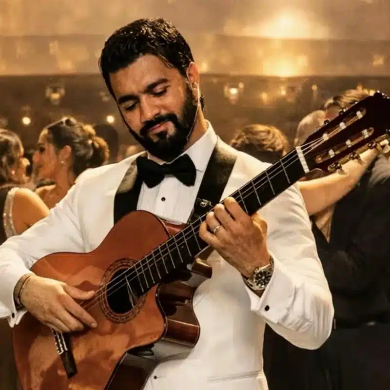Mouss, chanteur et guitariste live de l'équipe DJ Dadicool, en smoking blanc et nœud papillon jouant une guitare classique dans une salle de mariage à l'éclairage doré