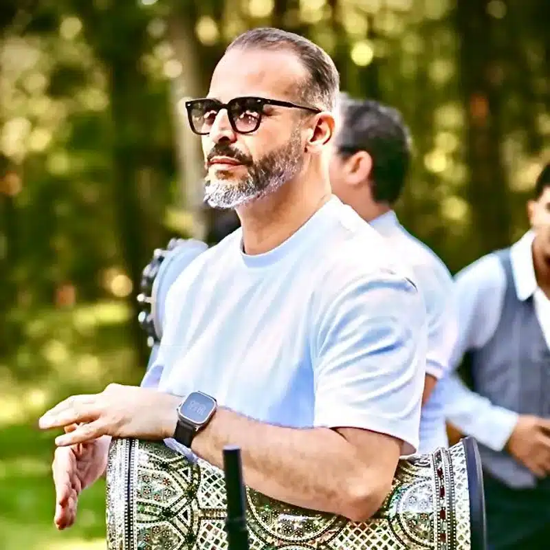 Ryad, percussionniste de l'équipe DJ Dadicool, jouant de la derbouka décorée en extérieur dans un parc, lunettes noires et barbe poivre et sel