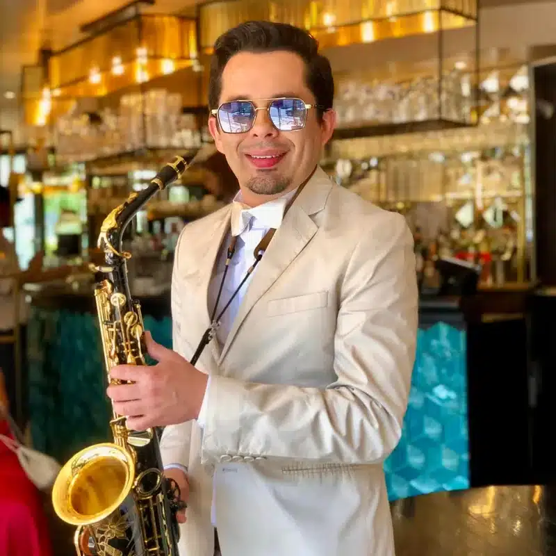 Saxo Gram, saxophoniste de l'équipe DJ Dadicool, portrait en costume crème, nœud papillon doré et lunettes de soleil aviator tenant son saxophone